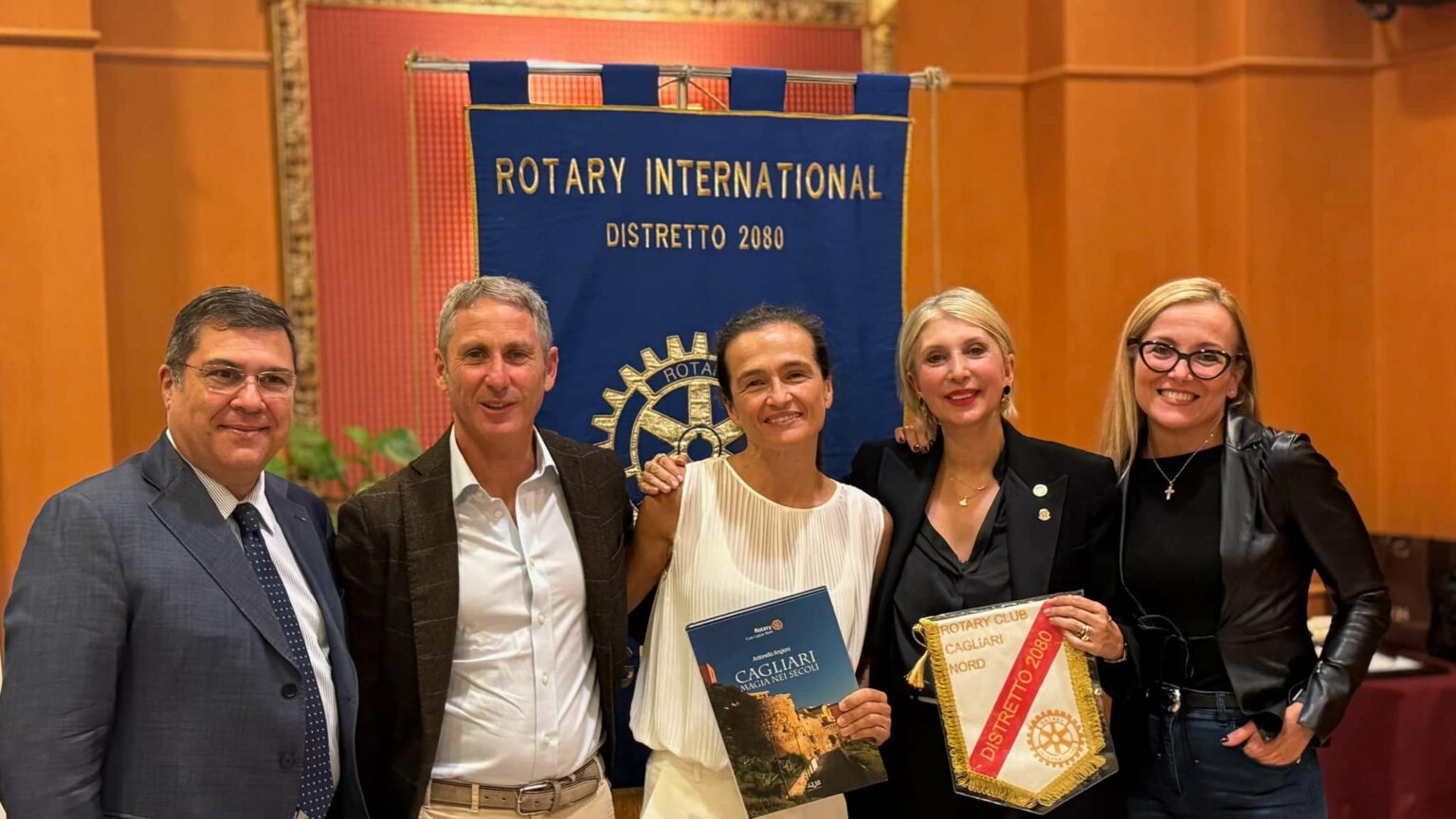 insieme di soci e socie del rotary club cagliari nord che tengono in mano il gagliardetto del club e sorridono guardando verso l'obiettivo